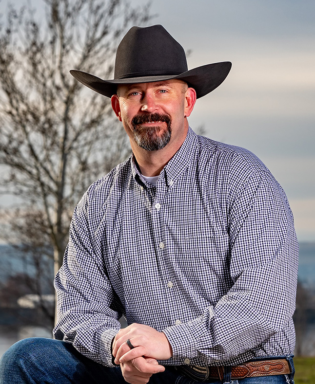 Chris Lee smiling with his hands crossed in a flannel and blue jeans with a cowboy hat